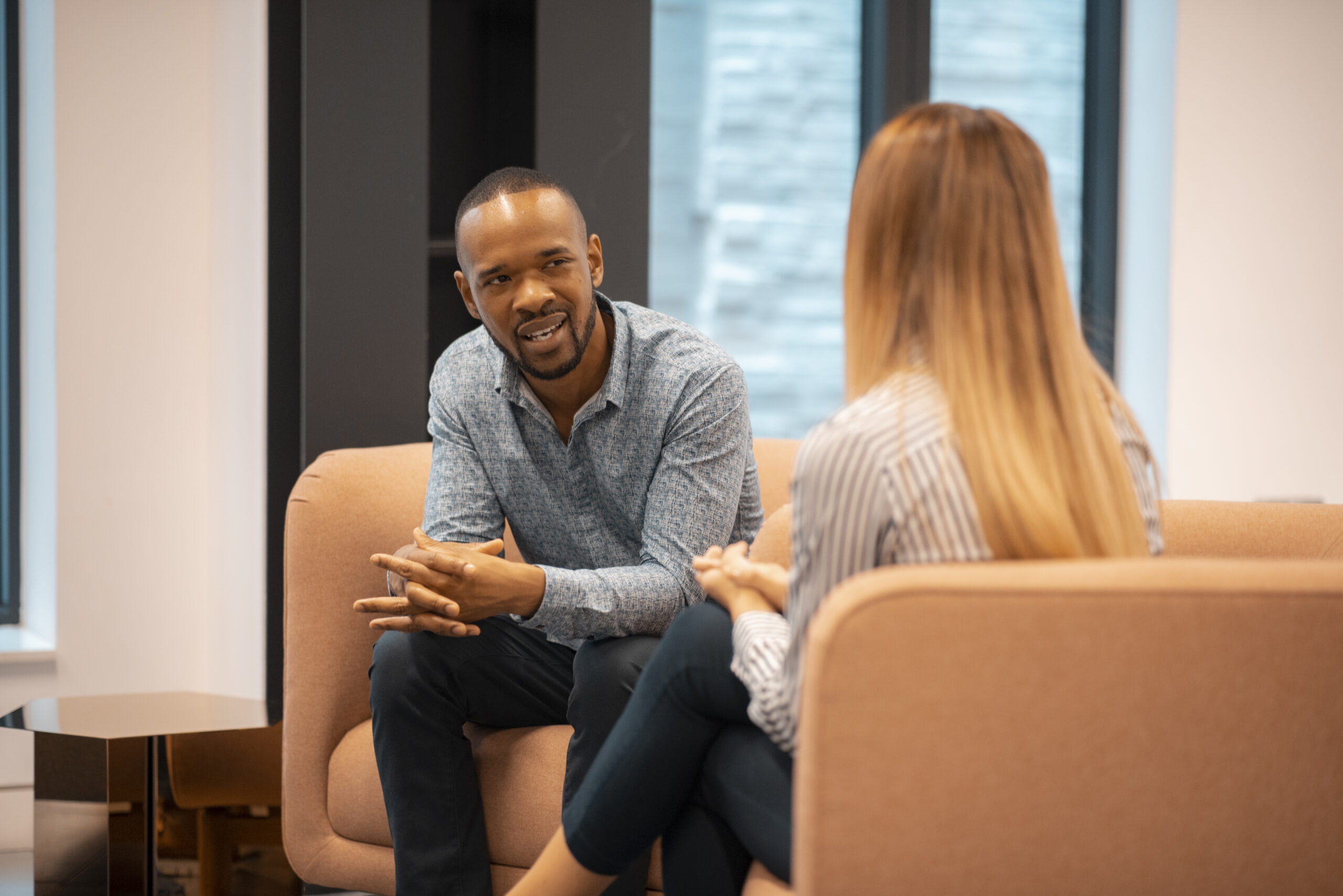 Photo of a man in a blue striped patterned shirt and trousers smiling in conversation with a woman with long blonde hair and a pinstriped shirt, on a beige sofa in a waiting area.
